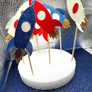 May include: A set of six rocket-shaped cake toppers, each with a different color and glitter finish. The toppers are made of felt and have wooden sticks for insertion into cakes. The toppers are arranged on a white foam cake base.