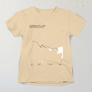 May include: A beige t-shirt with a black and white outline of Garibaldi Lake in Garibaldi Provincial Park, BC. The text "GARIBALDI LAKE" and "GARIBALDI PROVINCIAL PARK, BC" is printed on the shirt. The shirt also includes the text "8.2 km", "579 m", "6h 30 min", and "HARD".