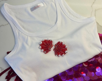 Puede incluir: Camiseta sin mangas blanca acanalada con dos aplicaciones de conchas rojas adornadas con perlas y cristales. La camiseta se combina con una falda de lentejuelas en tonos rosa y morado. La etiqueta de la camiseta sin mangas dice "LOVEROSE".