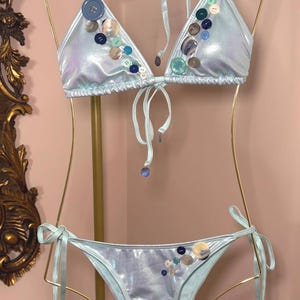 May include: A light blue bikini set with a metallic sheen, decorated with buttons of various sizes and colours. The triangle top and bottoms have tie closures. The bikini is displayed on a gold-coloured hanger.