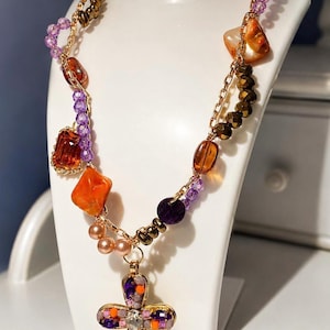Könnte beinhalten: Mehrreihige Halskette mit goldener Kette und Kreuzanhänger. Die Halskette hat orangefarbene, violette und goldfarbene Perlen. Der Kreuzanhänger ist mit farbigen Steinen und einem zentralen, klaren Edelstein verziert.
