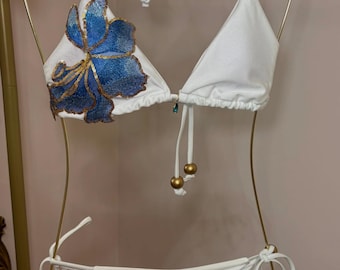 May include: White bikini set with blue floral embroidery. The triangle top and low-rise bottoms feature a delicate design with gold accents. The bikini is displayed on a gold hanger, showcasing its elegant details.