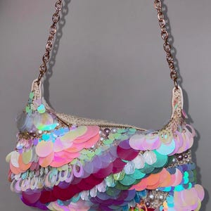 May include: A small, iridescent sequin handbag with a gold chain strap. The bag is decorated with a variety of colourful sequins, including pink, purple, green, and orange. Small white pearls are also visible along the top edge of the bag.
