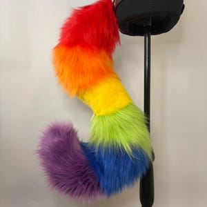 Könnte beinhalten: Ein regenbogenfarbener Kunstfellschwanz mit leuchtenden Farbtönen von Rot, Orange, Gelb, Grün, Blau und Lila. Der Schwanz ist flauschig und gebogen, an einer schwarzen Basis befestigt. Ein Kostümzubehör.