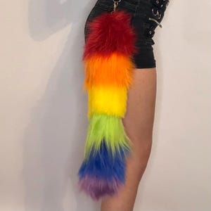 Puede incluir: Una cola de piel sintética de color arcoíris con un llavero. La cola está rayada verticalmente con rojo, naranja, amarillo, verde, azul y morado. La cola esponjosa está unida a un cierre de metal plateado.