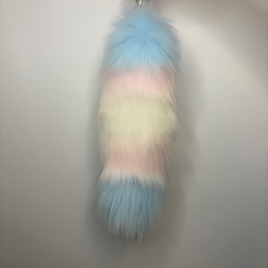 May include: A long, fluffy faux fur tail accessory featuring horizontal stripes in pastel blue, soft pink, and creamy white. The tail is tapered toward the bottom and includes a silver-toned metal clasp at the top for attachment, showcasing a thick, plush texture throughout the entire length of the item.