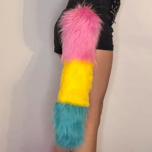 May include: A vibrant, fluffy tail accessory. The tail is segmented into three colours: pink at the top, yellow in the middle, and turquoise at the bottom. It is attached to a silver chain.