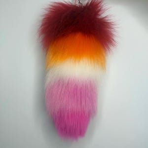 May include: A fluffy, long, tapered tail accessory made of faux fur. The tail features a horizontal gradient color pattern starting with deep red at the top, transitioning into bright orange, then white, and ending in a vibrant pink at the tip.