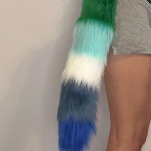 May include: A long, plush faux fur tail featuring a horizontal gradient pattern. The colours transition from a deep forest green at the top, through light turquoise, white, and slate grey, ending in a vibrant royal blue at the pointed tip.