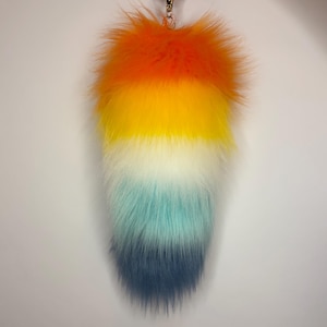 May include: A fluffy, faux fur tail accessory featuring a vibrant gradient color pattern. The colors transition vertically from a bright orange at the top, through yellow, white, and light turquoise, ending in a deep navy blue at the pointed base.
