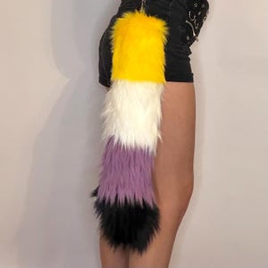 Puede incluir: Una cola con la bandera del orgullo no binario, con piel sintética amarilla, blanca, morada y negra. La cola está sujeta a una prenda negra. Un divertido accesorio para cosplay o para expresar la identidad.