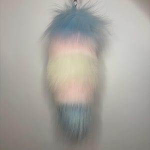 Puede incluir: Un accesorio largo y esponjoso de cola de piel sintética con un patrón de rayas horizontales en colores pastel. La secuencia de colores de arriba a abajo consiste en azul claro, rosa suave, blanco crema, rosa suave y azul claro, con un clip.