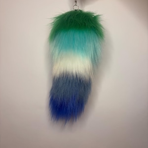 May include: A long, fluffy faux fur tail accessory featuring a vertical gradient colour pattern. The colours transition from a deep forest green at the top, through shades of teal and white in the middle, to a dark blue and bright royal blue at the tip.