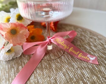 May include: A pink ribbon tied around a clear glass with the text "Kelsey's final fiesta" in gold lettering.