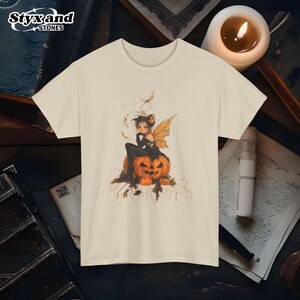 May include: A white t-shirt with a graphic of a fairy sitting on a jack-o'-lantern. The fairy has black wings and is wearing a black dress. The jack-o'-lantern is orange and has a carved face.