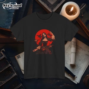 Puede incluir: Una camiseta negra con un estampado gráfico de una mujer con el pelo largo y negro sobre un fondo rojo. Ella sostiene cuerdas atadas a pájaros negros.