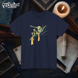 May include: A navy blue t-shirt with a graphic of a skeleton sitting on a branch. The skeleton is wearing a yellow scarf and has a skull head.
