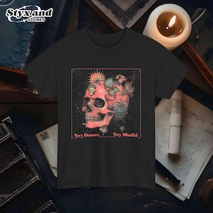 May include: Black t-shirt featuring a pink skull design with floral and botanical elements. The text "Very Demure, Very Mindful" is below the skull. The brand "Styx and Stones" is at the top left.