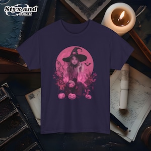 Puede incluir: Una camiseta morada con una ilustración rosa de una bruja con un sombrero puntiagudo y una luna llena detrás de ella. La bruja sostiene una calabaza y hay tres calabazas colgando de las ramas frente a ella.