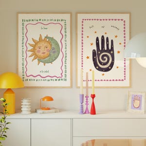 May include: Two framed wall art prints featuring celestial and mystical illustrations. The left print shows a sun and moon with the text "la lune et le soleil". The right print displays a dark hand with a spiral and the text "full of whimsy".
