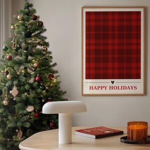 Op de afbeelding: Een kerstboom versierd met kerstballen en lichtjes staat naast een tafel met een witte lamp, een boek en een kaars. Een ingelijst print met een rood-zwart ruitpatroon en de tekst "Prettige Kerstdagen" hangt aan de muur.