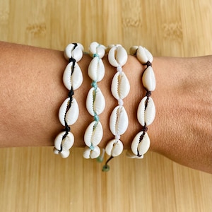 May include: Four shell bracelets with different coloured cords. The bracelets are made of white seashells and are tied with black, brown, white, and light blue cords.