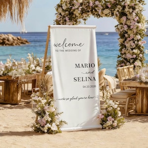 May include: A white wedding welcome sign on a wooden stand, with the words "welcome" and "Mario and Selina" in black script. The date 04.20.2026 is also displayed. The sign is decorated with flowers and set on a beach.