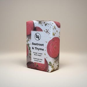 May include: A rectangular bar of handmade cold-pressed soap. The soap is wrapped in paper with a pattern of sliced beets and sprigs of thyme. The label reads "Beetroot & Thyme" with the ingredients: orange, juniper, and black pepper.