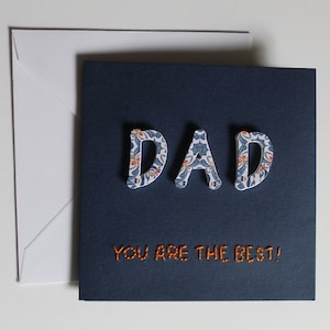 Handgemachte Karte zum Vatertag, ""DAD"" Wort in 3-D Schrift, gemustertes Papier auf dunkelblauer Karte."
