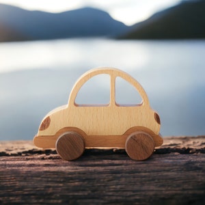 May include: A wooden toy car with a light brown finish. The car is sitting on a wooden surface with a blurred background of a lake and mountains.