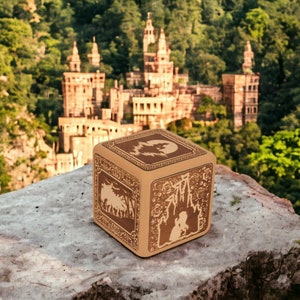 May include: A wooden box with intricate carvings depicting a forest scene, a castle, and a couple standing together. The box is sitting on a stone surface with a castle in the background.