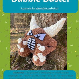 May include: A brown crocheted monster toy with white horns, eyes, and accents. The toy is sitting on a tree trunk. The image includes the text "Bubble Buster" and "A pattern by: @nerdyknotsbykari".