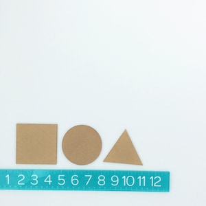 May include: Three brown cardboard shapes: a square, a circle, and a triangle, arranged from left to right on a white surface. A blue ruler with white numbers 1 through 12 is below the shapes.