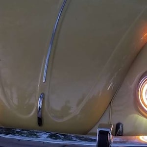 Może przedstawiać: Żółty zabytkowy Volkswagen Beetle z okrągłymi reflektorami i bursztynowymi kierunkowskazami. Reflektory są włączone, a samochód jest zaparkowany.