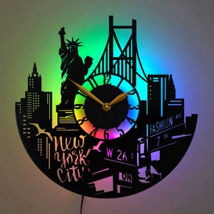 Puede incluir: Reloj de vinilo negro con una pantalla de luz LED de colores. El reloj presenta una silueta del horizonte de la ciudad de Nueva York, incluyendo la Estatua de la Libertad y el puente de Brooklyn. El reloj también incluye el texto "New York City" y señales de tráfico.