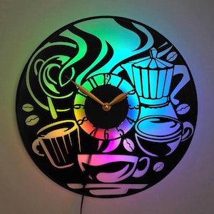 Op de afbeelding: Een zwarte vinyl platenspeler klok met een regenboogkleurig lichteffect. De klok heeft een koffiepot, koffiekopjes en stoom. De wijzers van de klok zijn goud.