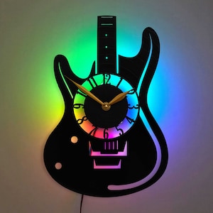 Puede incluir: Un reloj con forma de guitarra eléctrica negra con una esfera de colores arcoíris. Las manecillas del reloj son doradas y la guitarra tiene un fondo negro.