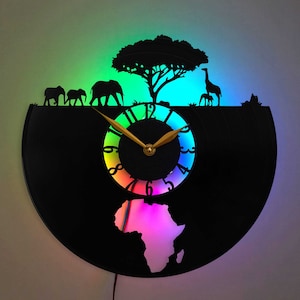Könnte beinhalten: Eine schwarze Schallplattenuhr mit einem regenbogenfarbenen Zifferblatt und einer Silhouette eines Baumes, Elefanten und einer Giraffe. Die Uhr wird mit einem Regenbogen an Farben beleuchtet, was einen lebendigen und farbenfrohen Effekt erzeugt.