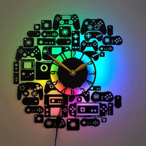 Op de afbeelding: Een zwarte en zilveren wandklok in de vorm van een videogamecontroller. De wijzerplaat van de klok is een regenboog van kleuren en heeft gouden wijzers. De klok is omgeven door verschillende vormen van videogamecontrollers.