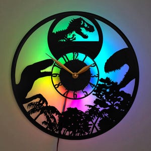 Puede incluir: Un reloj de vinilo negro con una pantalla LED de arcoíris. El reloj presenta una silueta de dinosaurios y árboles. La esfera del reloj tiene manecillas doradas y números negros.