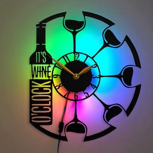 May include: Black wall clock with a wine bottle and glasses design. The clock face features the text "IT'S WINE O'CLOCK". The clock has gold-colored hands and is illuminated with a rainbow LED light. The clock is round and has a unique design.