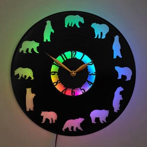 Puede incluir: Un reloj de vinilo negro con una esfera de reloj de color arcoíris y una silueta de osos alrededor del borde. El reloj tiene manecillas doradas y un centro dorado.