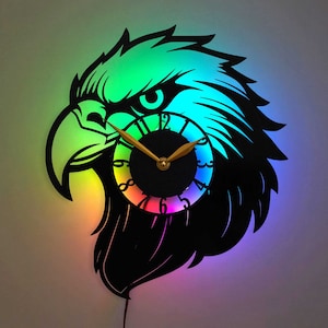 May include: A black eagle silhouette wall clock with a rainbow LED light effect. The clock face is round and features gold hands and a rainbow colored ring around the edge.