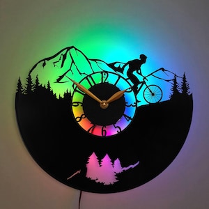Può includere: Orologio in vinile nero con silhouette di mountain biker, montagne e alberi. Il quadrante dell'orologio ha lancette dorate e un display luminoso a LED colorato. Un oggetto decorativo unico.