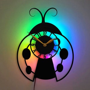 May include: A black ladybug-shaped clock with a rainbow-colored light behind it. The clock face has gold hands and black Roman numerals.