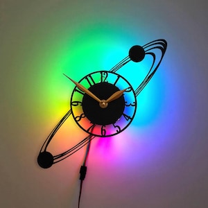 Peut inclure: Une horloge noire avec une lumière LED arc-en-ciel derrière elle. L'horloge est en forme de planète avec un anneau autour. Le cadran de l'horloge a des chiffres romains et des aiguilles dorées.