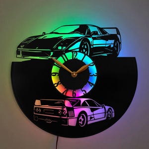 May include: A black vinyl record clock with a rainbow LED light display. The clock features a silhouette of a red Ferrari sports car on the top and bottom of the record. The clock hands are gold.