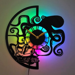 Op de afbeelding: Een zwarte octopusklok met een regenboogachtergrond. De wijzerplaat van de klok is wit met zwarte cijfers en gouden wijzers.