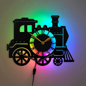 Puede incluir: Un reloj de metal negro con forma de tren con una esfera de colores arcoíris. El reloj tiene una aguja de hora y una de minutos de color dorado.