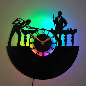 Könnte beinhalten: Eine schwarze Schallplattenuhr mit einem regenbogenfarbenen Zifferblatt. Die Uhr zeigt die Silhouette von zwei Personen beim Billardspielen. Die Uhr wird mit einem regenbogenfarbenen Licht beleuchtet.